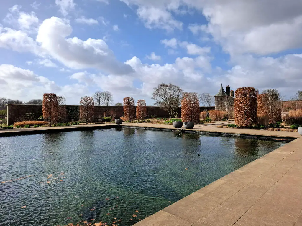 Calm reflective pool surrounded by tall, trimmed hedges and large potted plants under a sky with fluffy clouds. A historic building is visible in the distance.