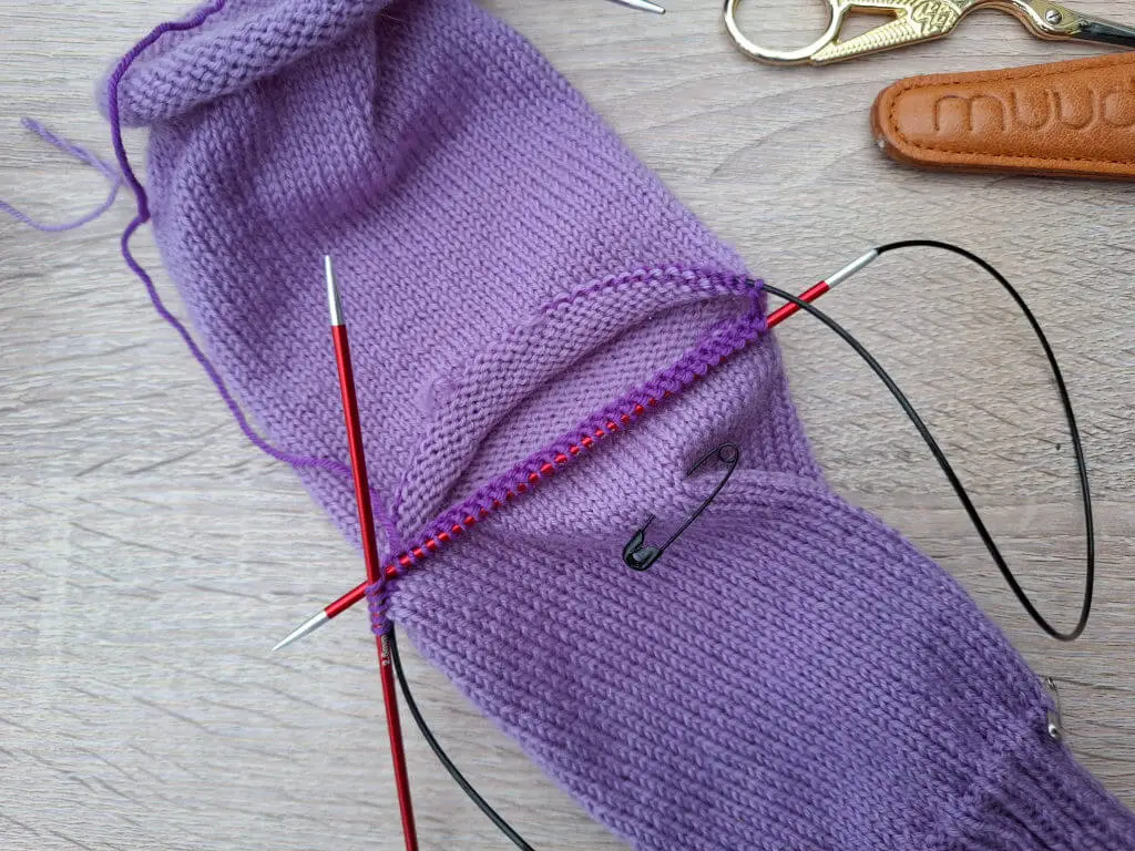 Knitting in progress with purple yarn on circular needles, forming part of a sock. A safety pin is attached. Scissors and a leather case lie nearby.