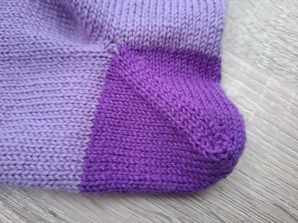 Close-up of a purple knitted sock on a wooden surface, showing the heel with light and dark purple sections, highlighting the texture and stitching.