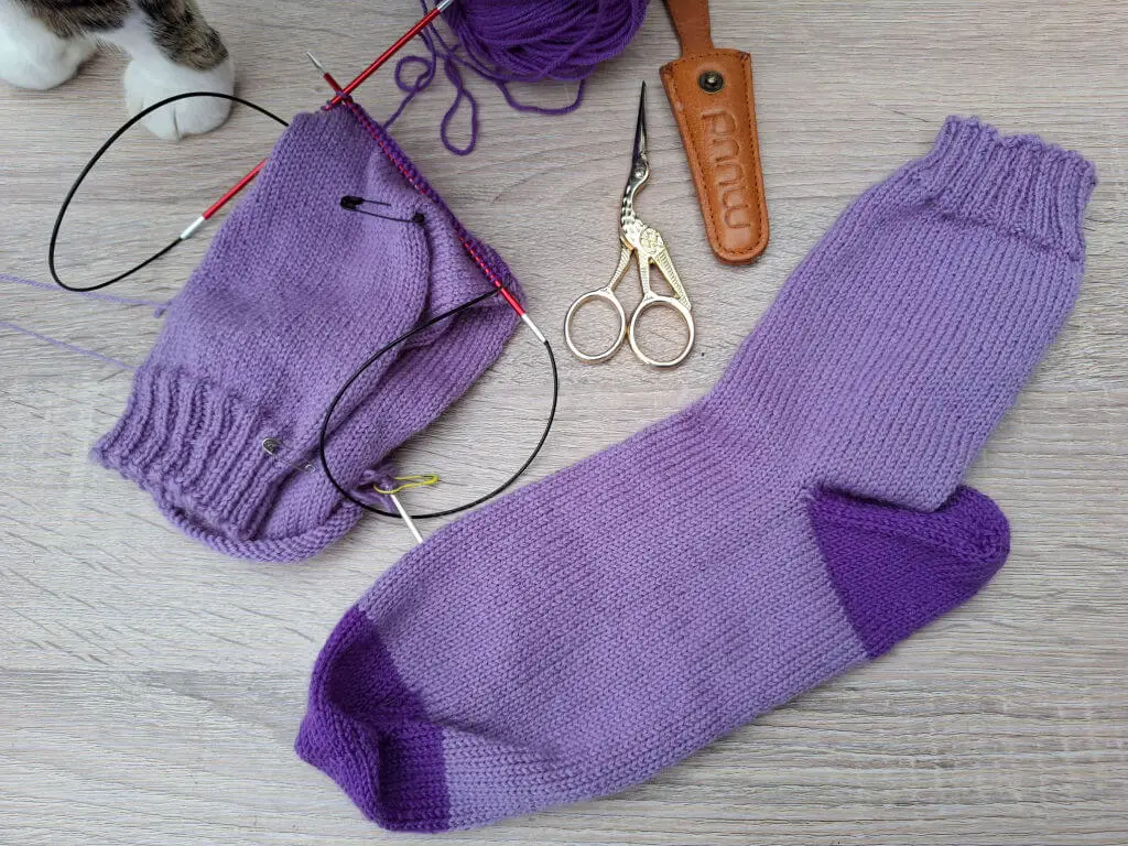 Purple knitted socks, one complete and one in progress on circular needles, lie on a wooden surface. Nearby are scissors, a leather sheath, and a ball of yarn. Partial view of a cat paw at the top left.