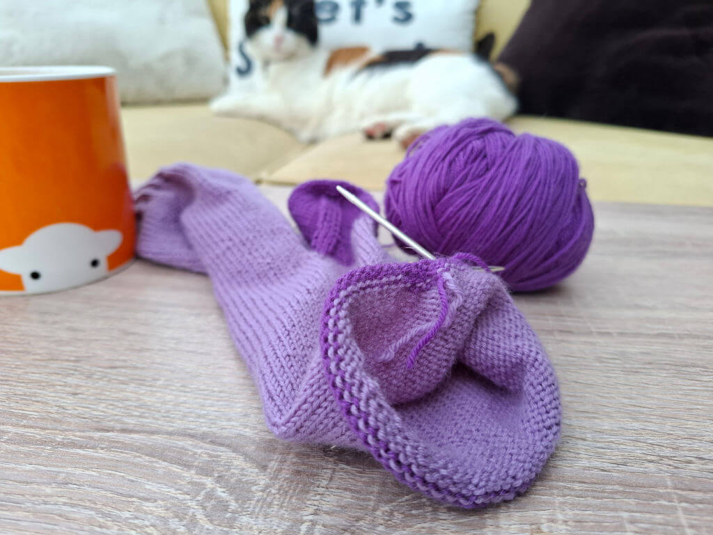 Purple knitting project with needles is on a wooden table. An orange mug with a sheep design is nearby. A cat lounges on a sofa in the background.