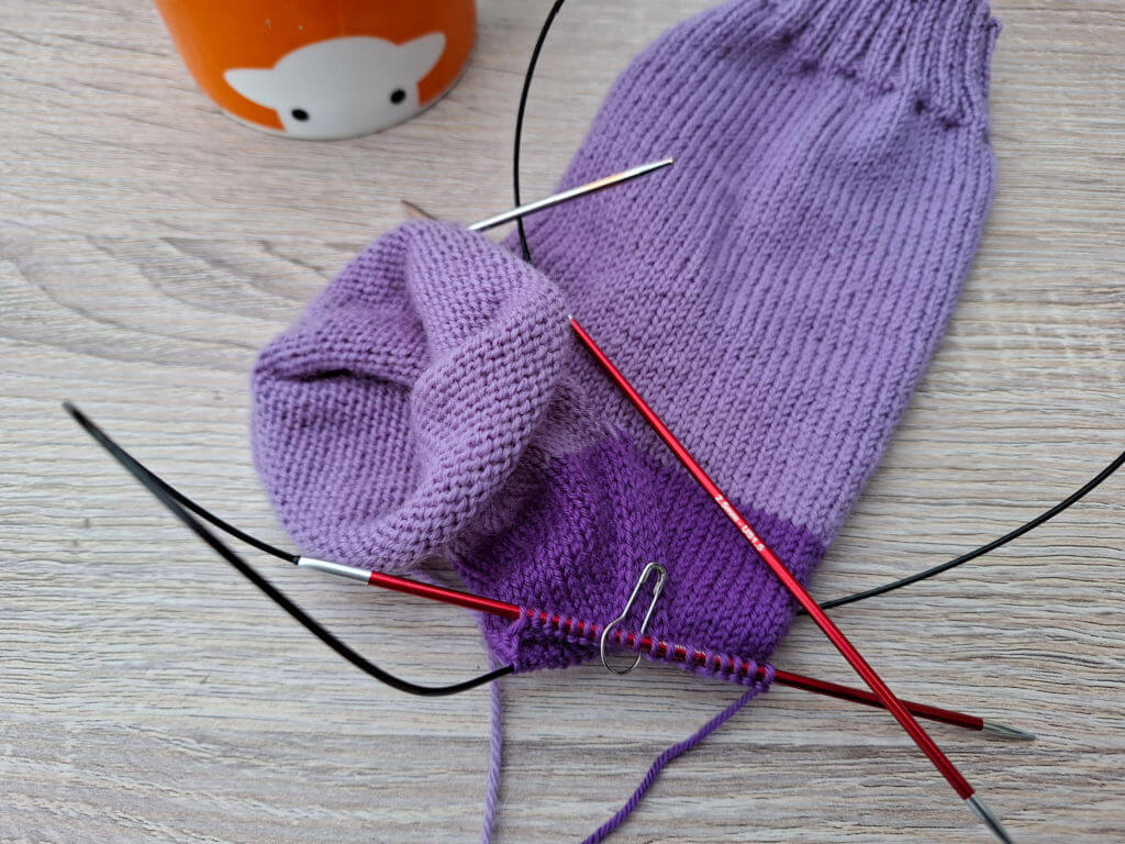 Close-up of a purple knit sock in progress with circular knitting needles. An orange mug with a sheep face is in the background on a wooden surface.