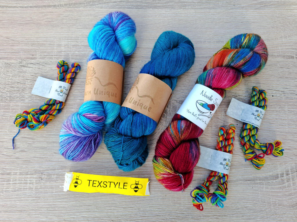 Various skeins of colourful yarn are arranged on a wooden surface. The yarns display vibrant hues of blue, red, purple, and multicoloured tones, conveying a creative and lively atmosphere. Bands with labels wrap around some skeins, and a yellow label reading "TEXSTYLE" is in the foreground.
