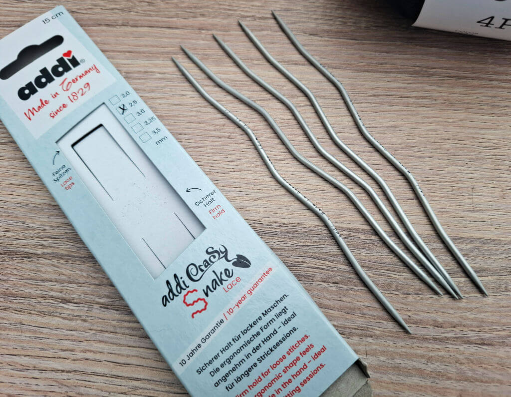 A set of five wavy metal knitting needles lie next to a light blue box labeled "addi CraSySnake" on a wooden surface, conveying a crafty, creative vibe.