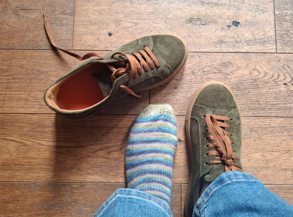 A foot in a colourful striped sock stands next to a pair of green suede sneakers with brown laces on a wooden floor, conveying a casual vibe.