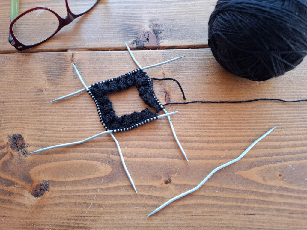 Knitting project on wooden table with black yarn and CrasySnake needles forming a square, next to a pair of glasses. Cosy and creative atmosphere.