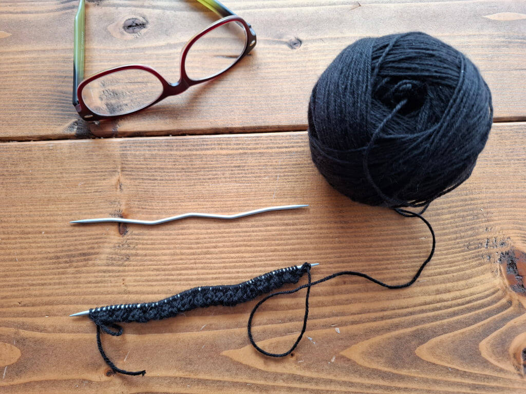 A skein of black yarn, red glasses, CrasySnake wavy knitting needles, and a partly knitted sock rest on a wooden table, creating a cosy crafting scene.