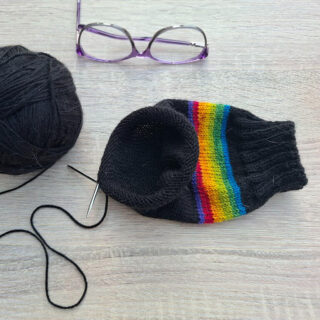 A ball of black yarn, partially knitted sock with rainbow stripes, a knitting needle, and purple glasses are on a light wooden table. Cosy crafting mood.