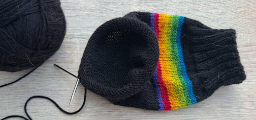 A ball of black yarn, partially knitted sock with rainbow stripes, a knitting needle, and purple glasses are on a light wooden table. Cosy crafting mood.