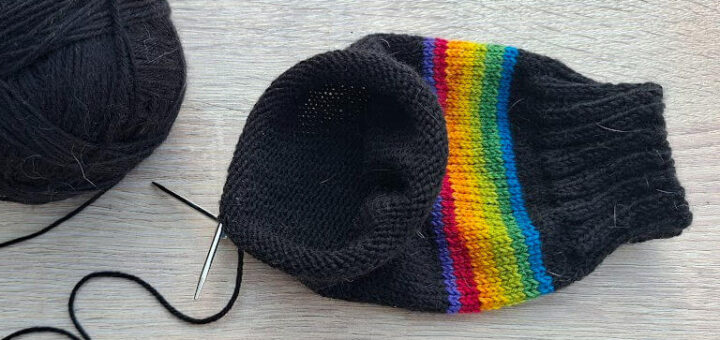 A ball of black yarn, partially knitted sock with rainbow stripes, a knitting needle, and purple glasses are on a light wooden table. Cosy crafting mood.