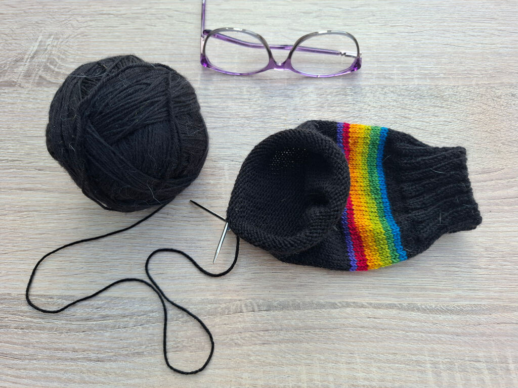 A ball of black yarn, partially knitted sock with rainbow stripes, a knitting needle, and purple glasses are on a light wooden table. Cosy crafting mood.