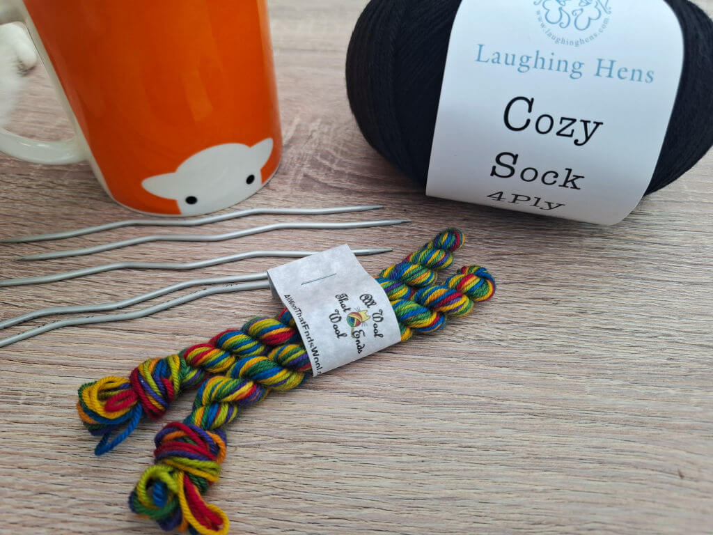 Orange sheep mug, black yarn labeled "Cozy Sock," colourful yarn micro mini skeins, and silver knitting needles on a wooden surface. Crafty, cosy vibe.