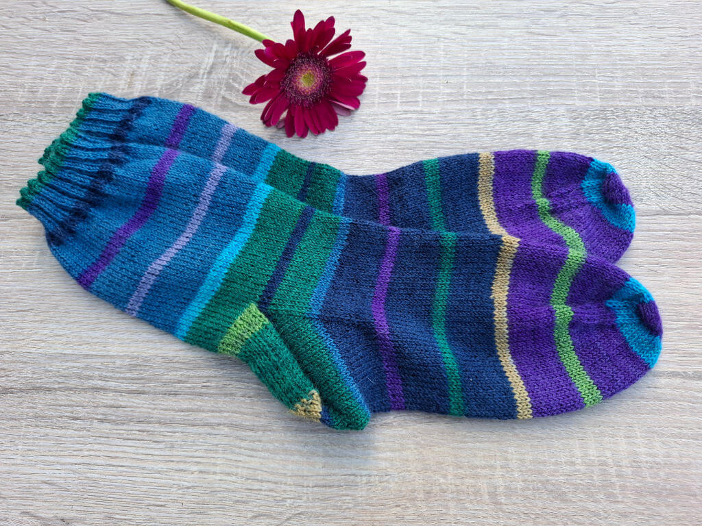 Colourful striped socks in shades of blue, purple, and green are neatly arranged on a light wooden surface with a vibrant red flower above them.