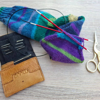 Knitting tools on a table: colourful striped sock in progress with circular needles, a brown needle case, and elegant gold scissors nearby.
