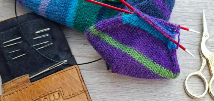 Knitting tools on a table: colourful striped sock in progress with circular needles, a brown needle case, and elegant gold scissors nearby.