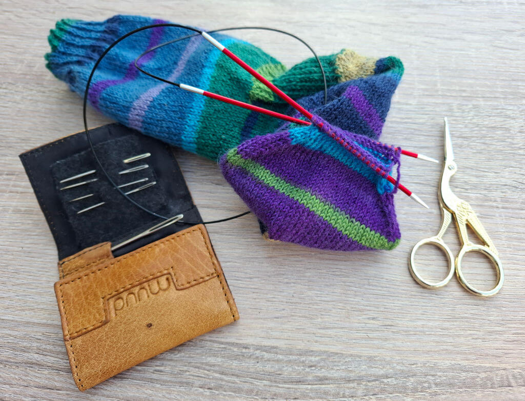 Knitting tools on a table: colourful striped sock in progress with circular needles, a brown needle case, and elegant gold scissors nearby.