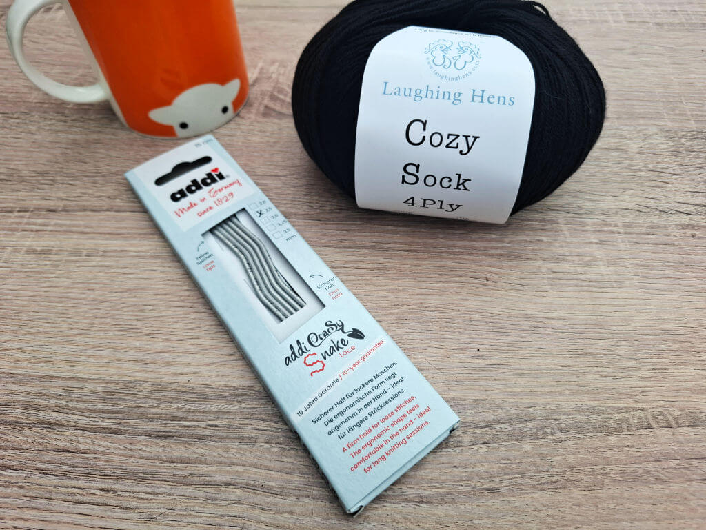 A ball of black "Cozy Sock" 4-ply yarn and a set of metal knitting needles are on a wooden surface. An orange mug with a sheep design is in the background.