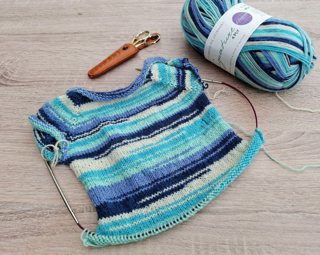Knitted striped fabric in blue and teal hues on circular needles; yarn ball and scissors lie nearby on a wooden table.
