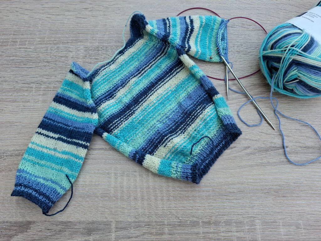 Knitting project with blue striped baby sweater, partially completed. Needles are attached, yarn ball beside, on a wooden surface. Cosy and crafty feel.