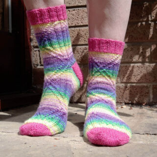 Feet wearing colourful, hand-knit socks with pink, green, blue, and purple stripes. The background is a textured brick wall. Bright and cheerful tone.