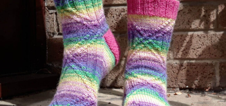 Feet wearing colourful, hand-knit socks with pink, green, blue, and purple stripes. The background is a textured brick wall. Bright and cheerful tone.