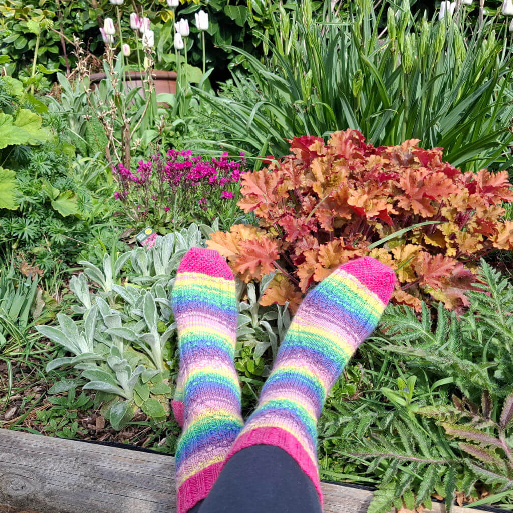Person with colourful, striped socks relaxes in a lush garden with diverse plants, including blooming flowers and vibrant foliage, under sunny skies.