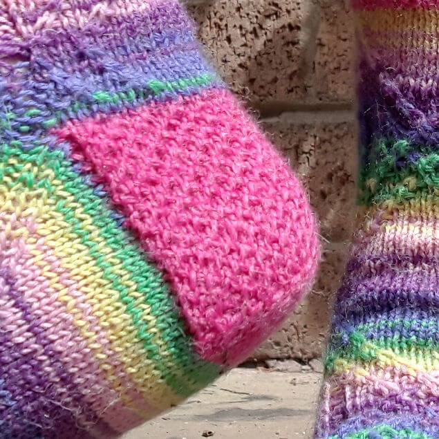 Close-up of colourful, knitted socks against a brick wall. One sock features a vibrant pink heel, while both display stripes in green, yellow, and purple. Cosy and cheerful.