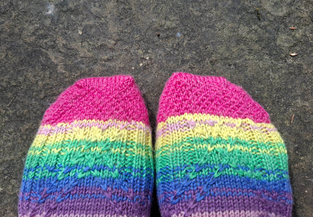 Two feet wearing bright, colourful, rainbow-striped knitted socks stand on a textured grey stone surface. The vibrant colours evoke a cheerful, playful mood. Winwick Mum | Kaleidoscope Socks