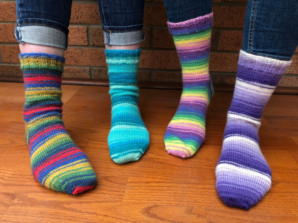 Four feet in colourful, striped socks in WYS Winwick Mum yarn on a wooden floor. Each sock displays a unique pattern and colour scheme, exuding warmth and cheerfulness.