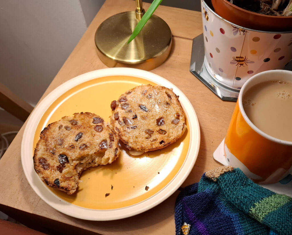 A cosy setting features buttery toasted hot cross bun studded with raisins on a yellow plate. Beside them is a cup of tea and a partly-knitted sock on a wooden table.