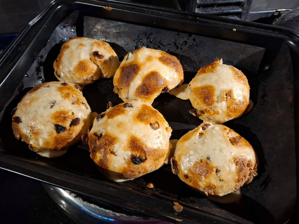 Six golden-brown hot cross buns with raisins rest on a dark baking tray. The buns are slightly glossy, indicating a fresh glaze.