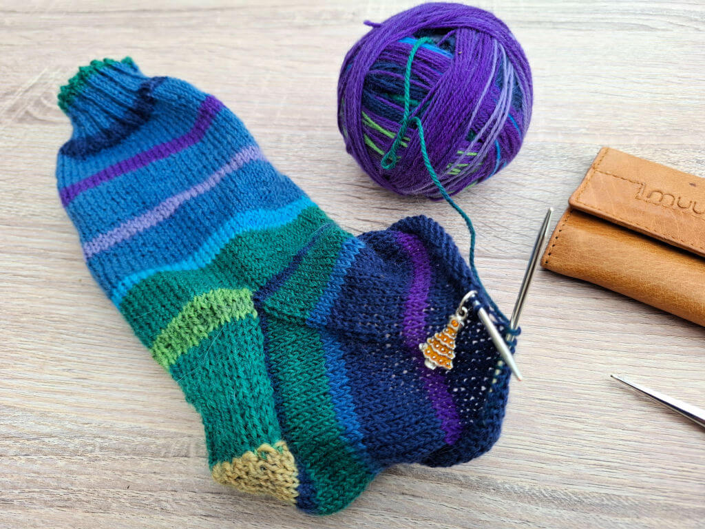 A colourful knitted sock in progress, featuring blue, green, and purple stripes, with a knitting charm. A ball of purple yarn and knitting needles are beside it.