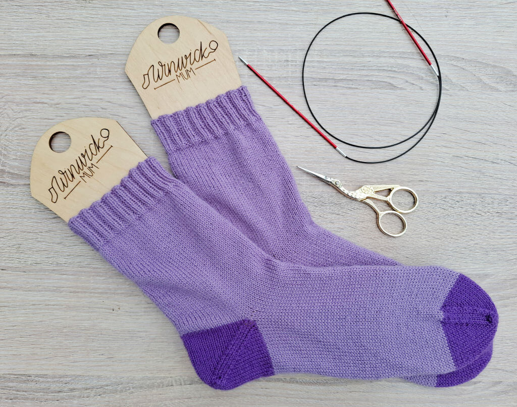 Hand-knitted purple socks with darker purple toes on wooden sock blockers, surrounded by a circular knitting needle and vintage-style scissors on a light wooden surface.