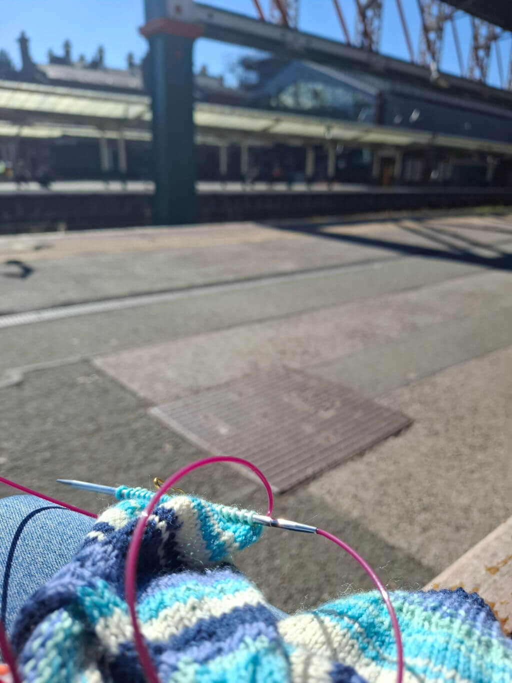 Person knitting with blue and white yarn at a sunny train station. Blurred tracks and platform canopy in the background evoke a calm atmosphere.