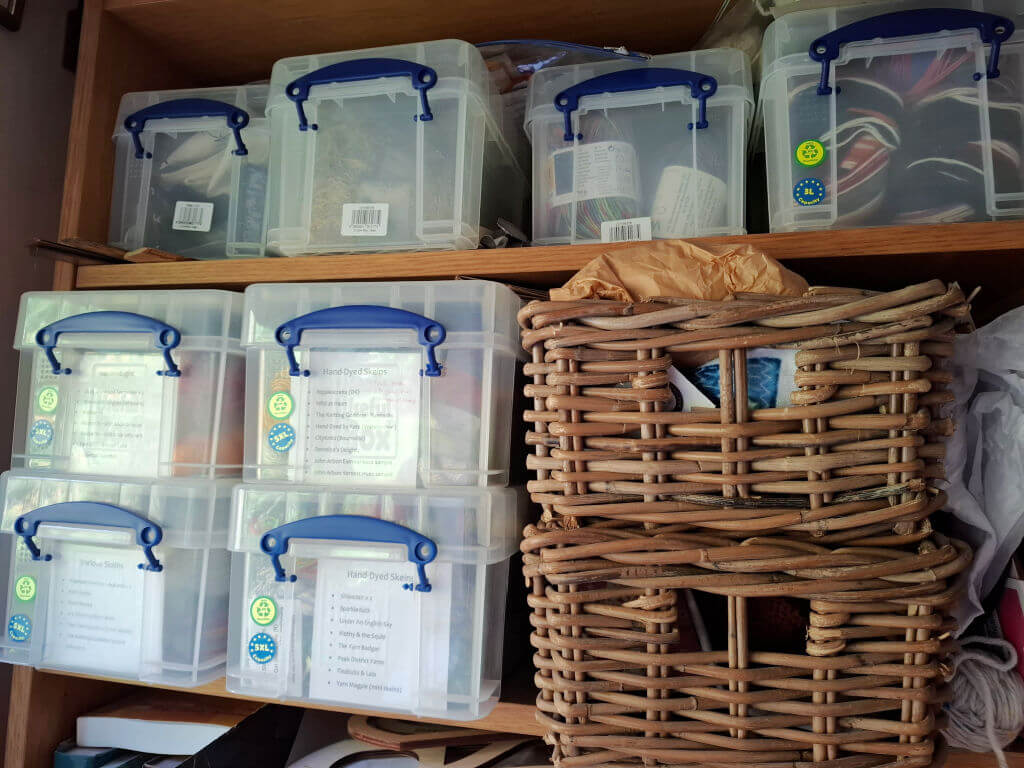 Plastic storage boxes with blue handles organised on shelves, labelled with stickers, alongside two wicker baskets. The scene feels tidy and organised.