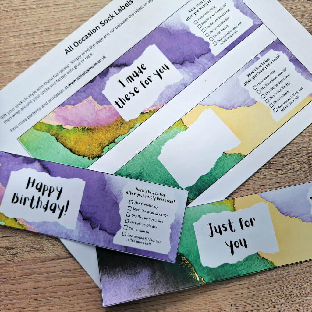 Colourful sock labels on a wooden surface, featuring watercolour backgrounds with text like "Happy Birthday" and "Just for You." Labels include care instructions.