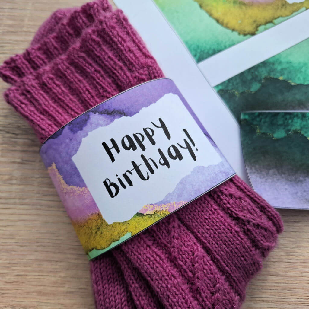 Knitted pink socks with a "Happy Birthday!" tag featuring a colourful watercolor design, placed on a wooden surface with other labels nearby.