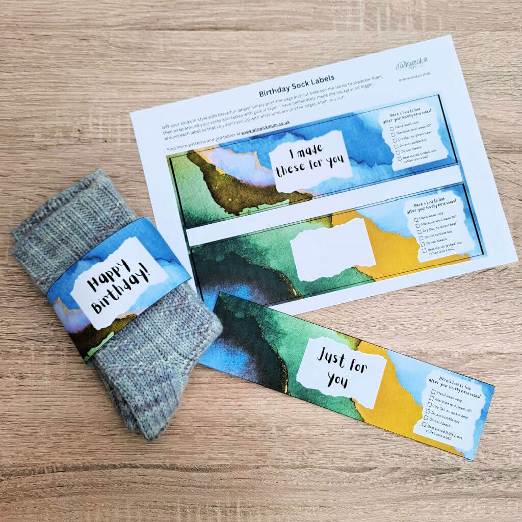 A handmade sock gift set on a wood surface with colourful watercolour labels. One label wraps blue socks with "Happy birthday!" text; others read "I made these for you" and "Just for you."