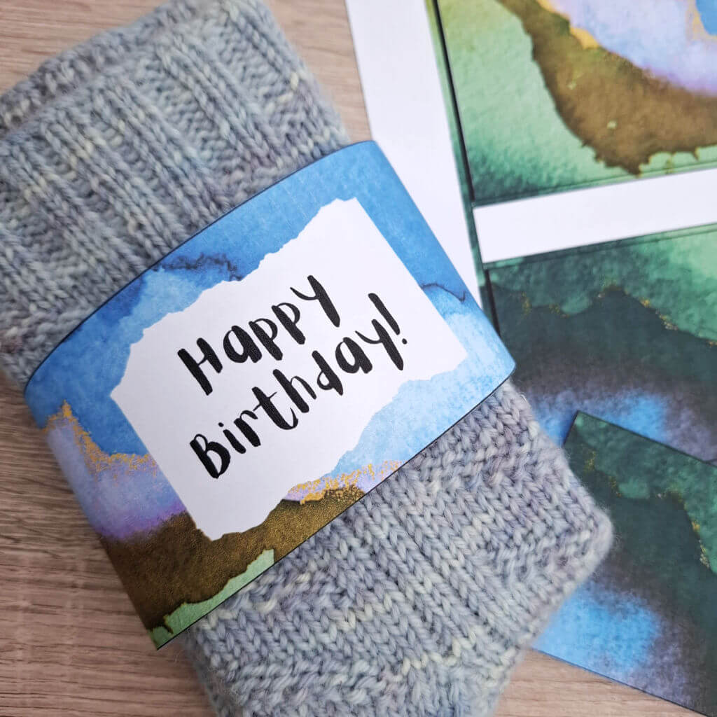 Close-up of a folded blue knit sock with a colourful, abstract band labeled "Happy Birthday!" in bold text. The tone is warm and festive.
