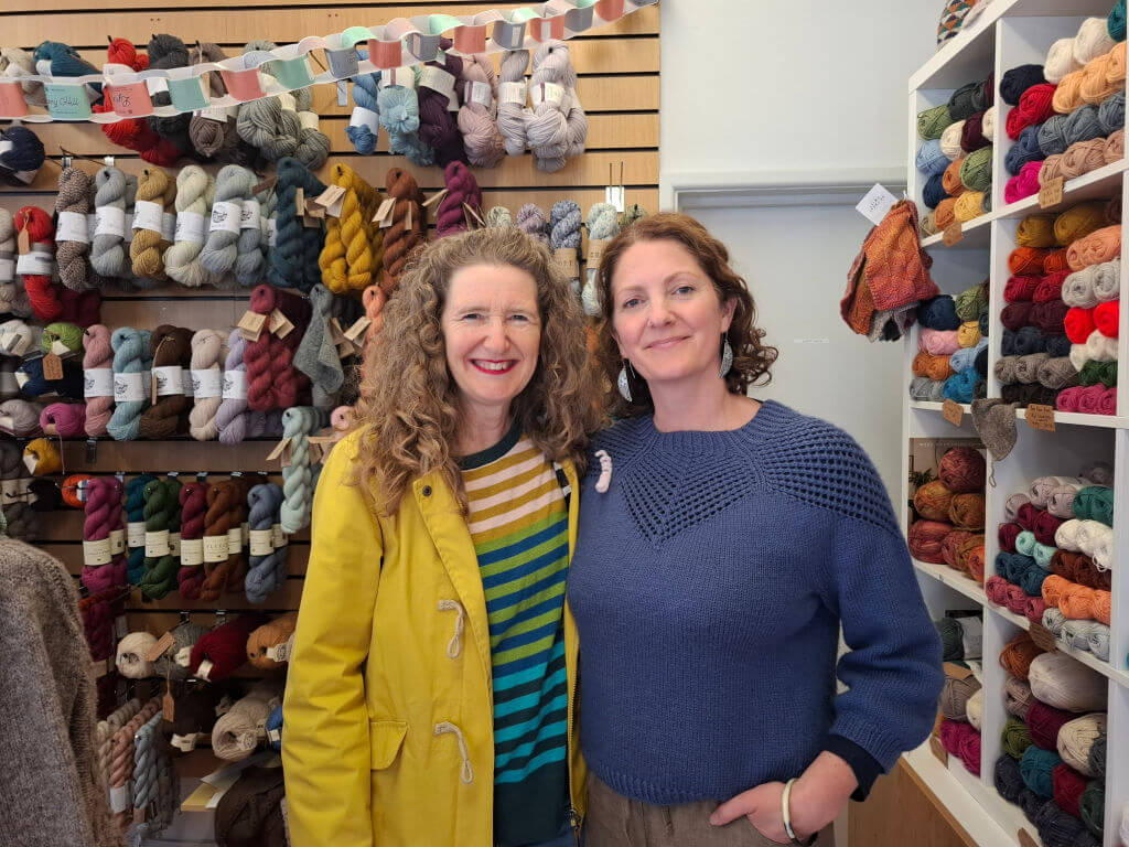 Two women smile warmly in a vibrant yarn shop. Shelves are stocked with colourful yarn, creating a cosy, creative atmosphere. The mood is cheerful and inviting.