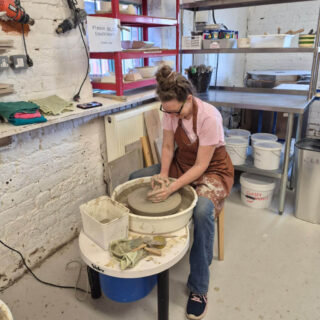 A woman in a pink shirt and apron is focused on shaping clay on a pottery wheel in a workshop. The room has shelves with tools and materials.