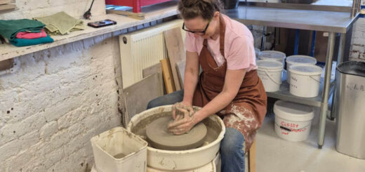 A woman in a pink shirt and apron is focused on shaping clay on a pottery wheel in a workshop. The room has shelves with tools and materials.