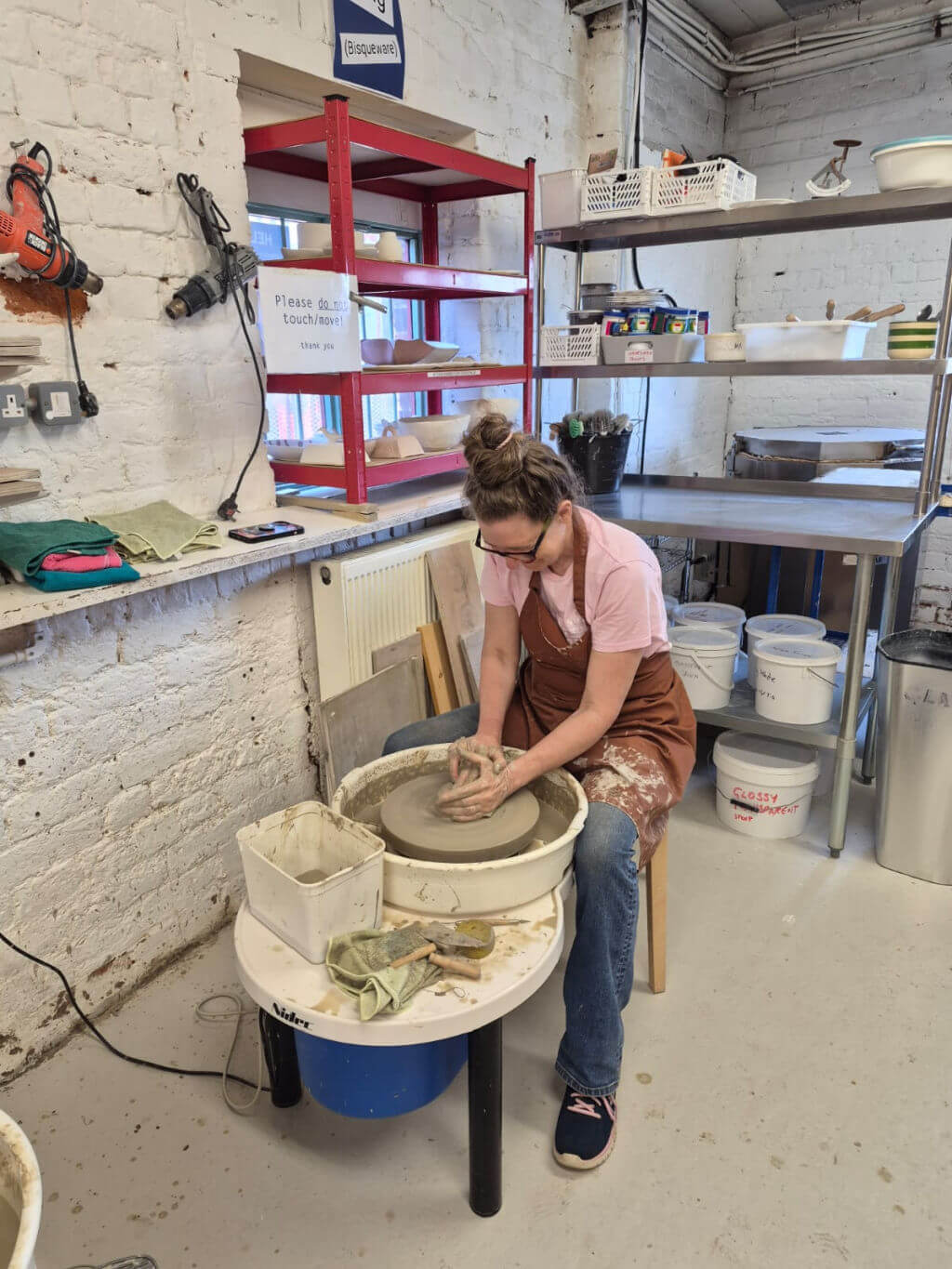 A woman in a pink shirt and apron is focused on shaping clay on a pottery wheel in a workshop. The room has shelves with tools and materials.