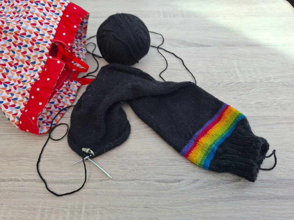 A colourful knitting bag sits on a wooden table beside a black knitted piece with a rainbow stripe and attached knitting needles, evoking creativity.