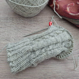 A partially knitted grey sock with intricate cable patterns on wooden surface, featuring a metal knitting needle and red stitch marker. A yarn ball and fabric in the background convey a cosy atmosphere.