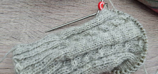 A partially knitted grey sock with intricate cable patterns on wooden surface, featuring a metal knitting needle and red stitch marker. A yarn ball and fabric in the background convey a cosy atmosphere.