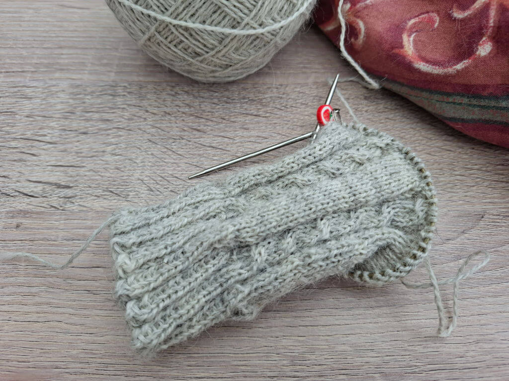 A partially knitted grey sock with intricate cable patterns on wooden surface, featuring a metal knitting needle and red stitch marker. A yarn ball and fabric in the background convey a cosy atmosphere.