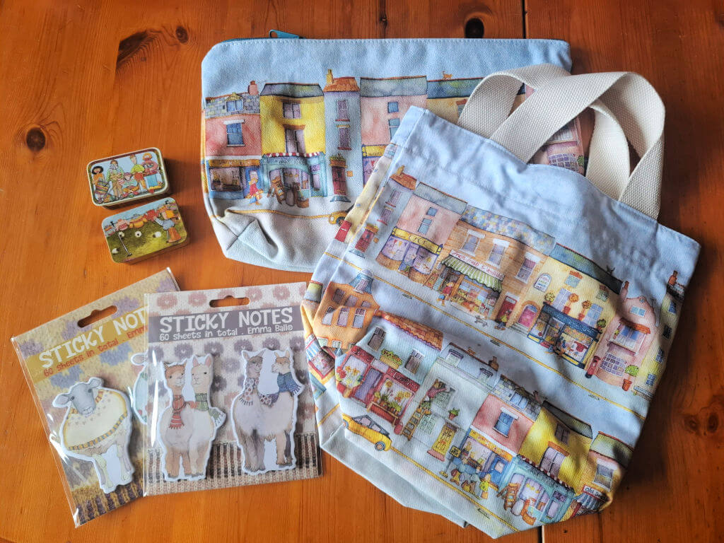 Tote bag, knitting pouch, and tins featuring colourful town illustrations on a wooden table. Two packs of novelty sticky notes with whimsical animal designs.