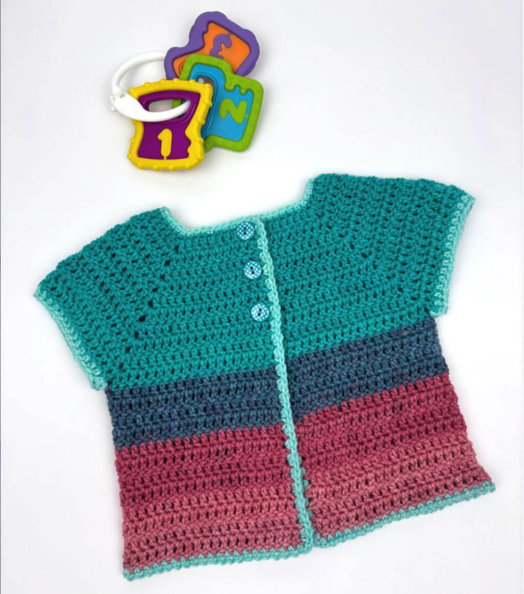 Crocheted baby cardigan in teal, blue, and pink, with three light blue buttons. Nearby are colourful number toy links on a white surface.