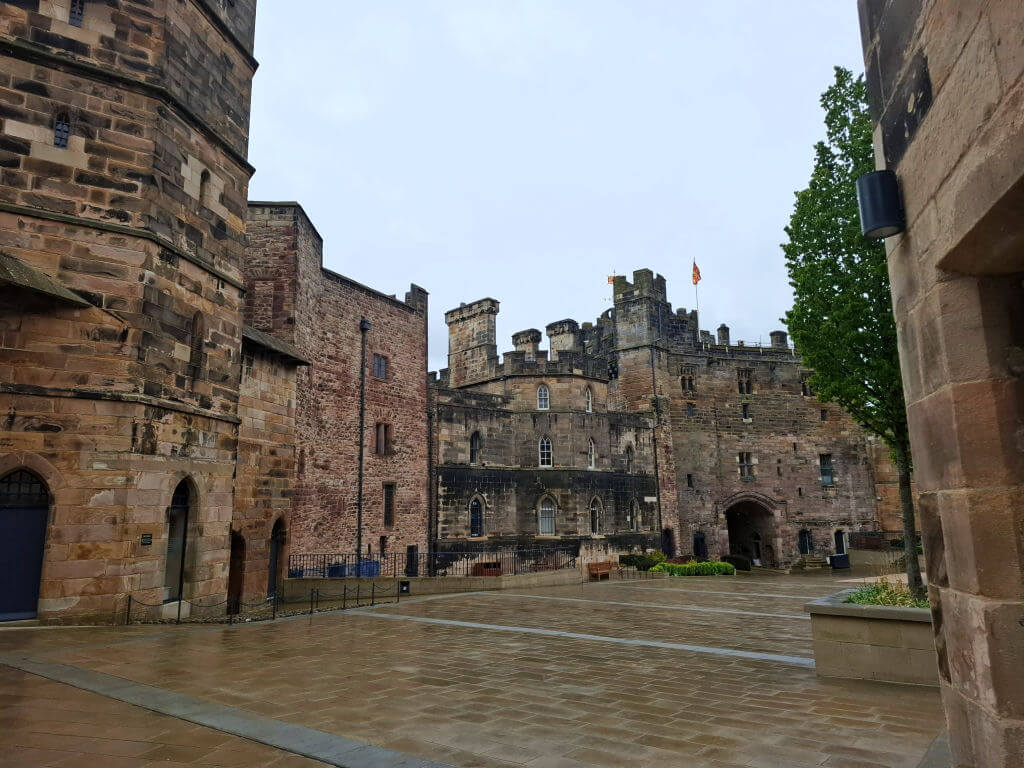 Medieval castle courtyard with stone buildings and tall towers, featuring a flag on one tower. Wet cobblestone reflects a rainy, overcast atmosphere.